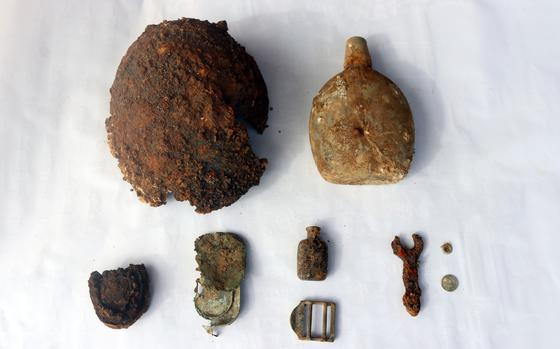 A collection of rusted and corroded wartime artifacts displayed on a white surface, including a damaged helmet, a canteen, small bottles, a buckle, a wrench and various metal fragments.