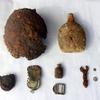 A collection of rusted and corroded wartime artifacts displayed on a white surface, including a damaged helmet, a canteen, small bottles, a buckle, a wrench and various metal fragments.