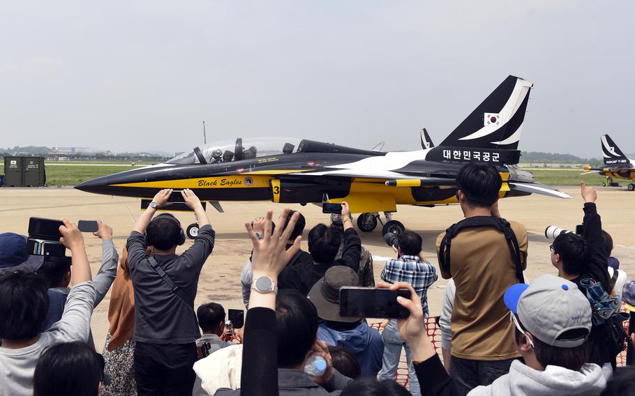 A military fighter jet taxis on a runway as a crowd of people, seen from behind, use phones to take photos.