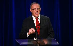 A man in a suit and red tie speaks vigorously at a podium.