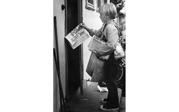 A boy stands in front of a door carrying newspapers.