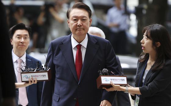 A South Korean man in a dark suit and red tie walks forward.