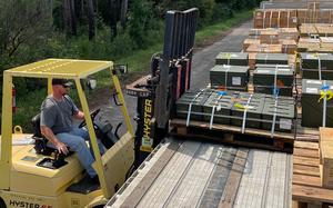 A man operates a yellow forklift to move pallets of military ammunition crates onto a flatbed truck. 