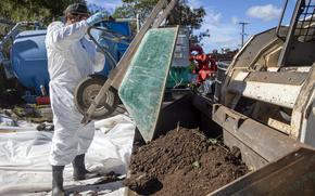 A man in a white hazmat suit dumps soil from a wheelbarrow into a large scoop attached to a tractor.