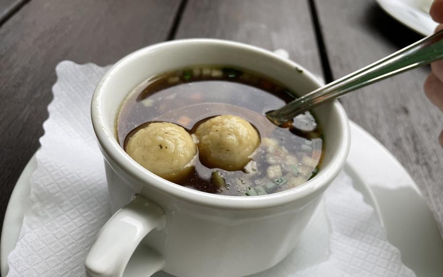 A close-up of a broth soup served in a white ceramic mug, containing two round dumplings and garnished with fresh herbs. A spoon rests in the soup, and the dish is served on a white napkin on a wooden table surface.