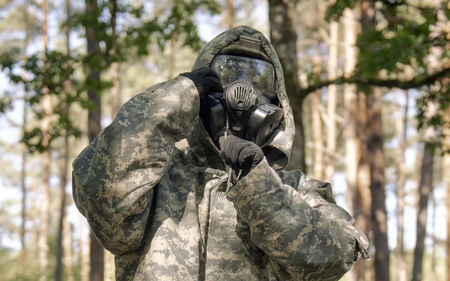 A soldier in full gear, pulls a hood over his head and gas mask.