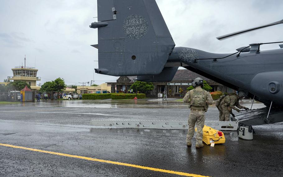 Two military service members in camouflage flights suits and helmets stand by the open rear hatch at the back of a cargo plane on a runway.