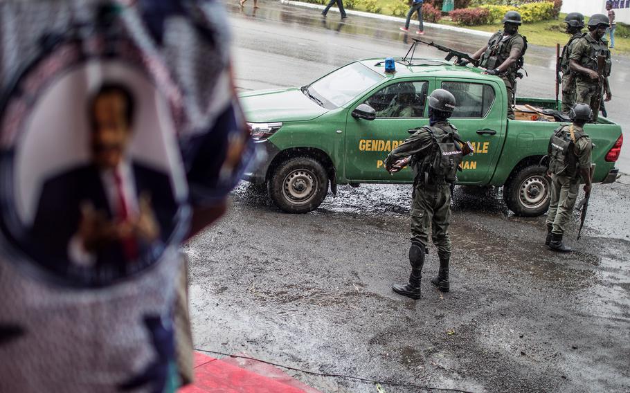 A supporter of the ruling CPDM party, Cameroon People’s Democratic Movement of incumbent Cameroonian President Paul Biya, wears a shirt emblazoned with the pictures of the President as a patrol of the Cameroonian Gendarmerie deploys in the Omar Bongo Square of the majority anglophone South West province capital Buea, on Oct. 3, 2018, on the sidelines of a political rally.