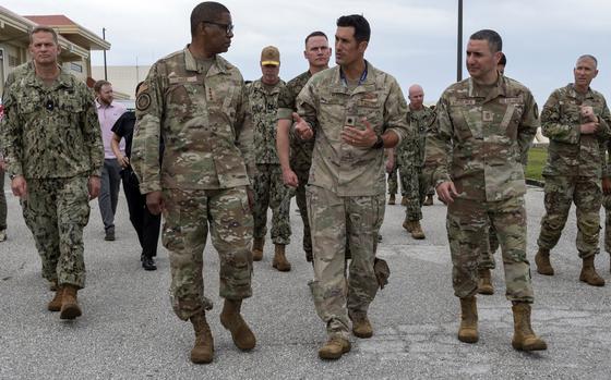 Military personnel from multiple service branches walk together on a paved surface at a military installation. The group includes officers in various camouflage uniforms. Several are engaged in conversation as they walk, with buildings and overcast sky visible in the background. 
