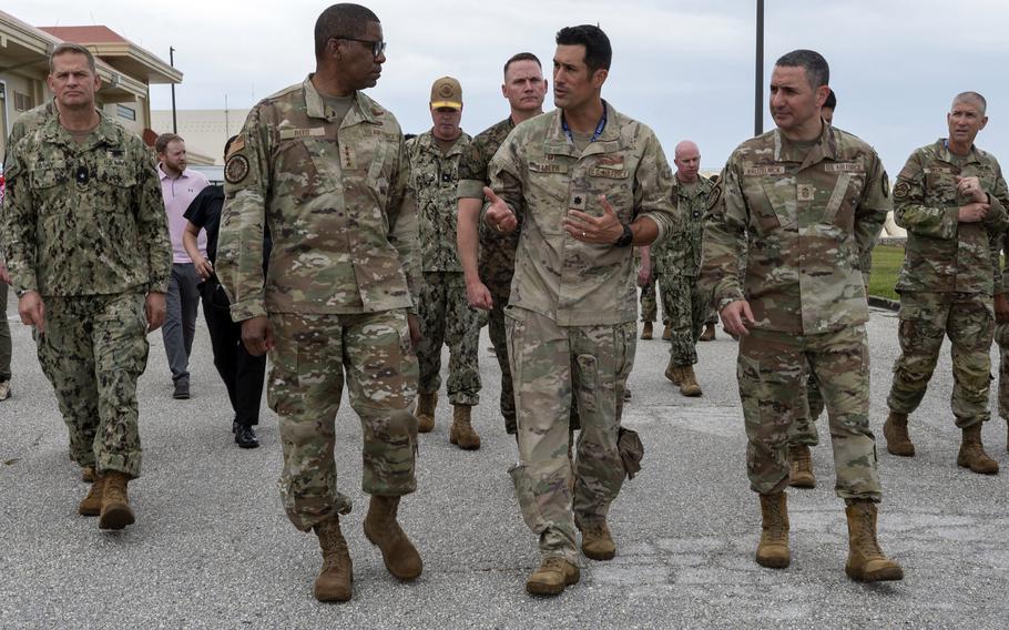Military personnel from multiple service branches walk together on a paved surface at a military installation. The group includes officers in various camouflage uniforms. Several are engaged in conversation as they walk, with buildings and overcast sky visible in the background. 