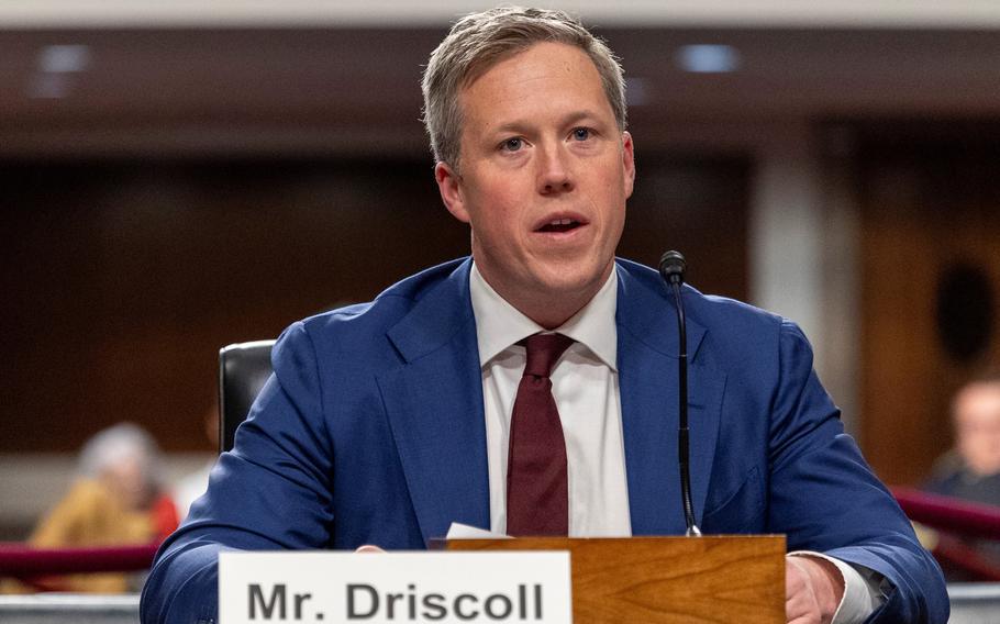 A man in a suit sitting at a table and speaking.