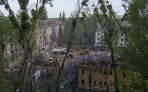 Construction vehicles, seend from a distance through tree branches, clear rubble and debris from a square between damaged buildings.