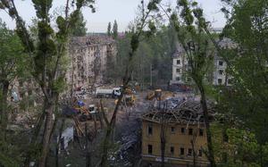 Construction vehicles, seend from a distance through tree branches, clear rubble and debris from a square between damaged buildings.