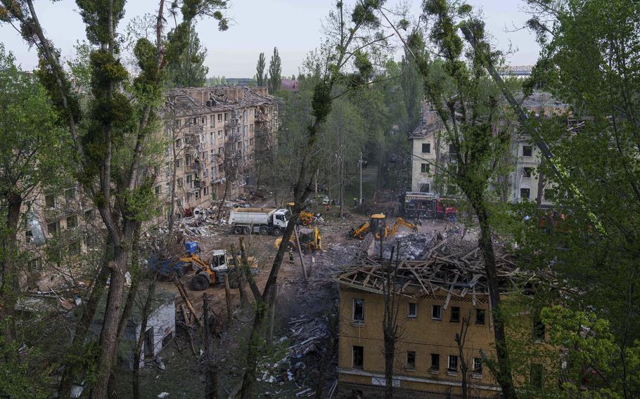 Construction vehicles, seend from a distance through tree branches, clear rubble and debris from a square between damaged buildings.
