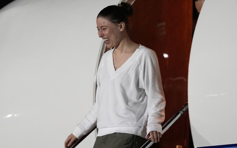 A woman in a white sweater smiles as she stands at the top of a staircase while exiting an airplane.