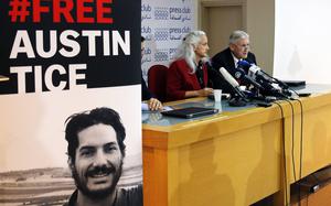 A large poster with a man’s photo reading “#Free Austin Tice” stands next to a table where the man’s parents sit and speak into a row of microphones.