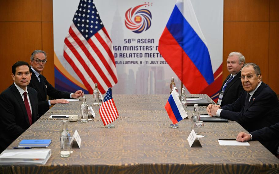 American and Russian officials sit on opposite sides of a conference table, with small U.S. and Russian flags on the table in front of them and large flags behind.