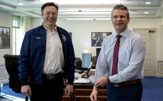 Elon Musk, wearing a dark blue jacket with his last name embroidered on a name tape, stands next to Defense Secretary Pete Hegseth. 