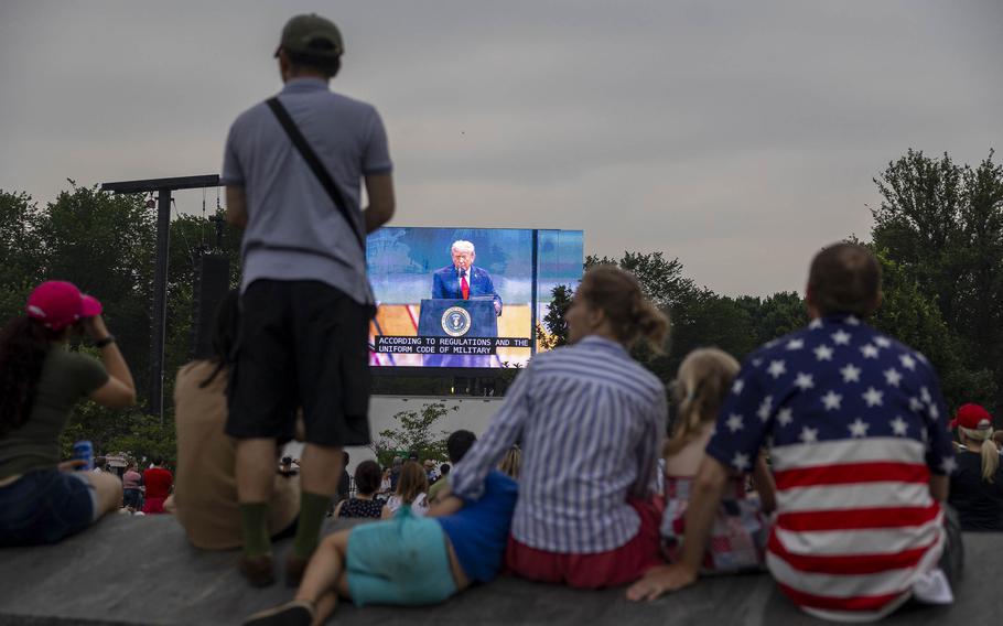 A crowd of people sitting outside looking at a screen with Donald Trump on it.