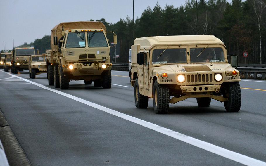 A military convoy travels on a paved highway. In the foreground, a tan-colored Humvee leads the formation with its headlights on. Behind it, a larger military truck and additional vehicles follow in single file. 