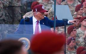 Donald Trump, wearing a baseball cap, points to his left as he gives a speech surrounding by soldiers in camouflage uniforms.