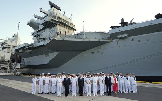 A group of British sailors in white uniforms and officials in suits pose on a dock in front of an aircraft carrier.
