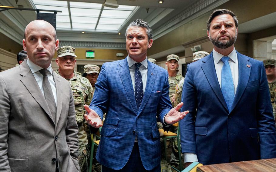 Stephen Miller, Pete Hegseth and JD Vance stand at a table in front of National Guard members in camouflage uniforms.