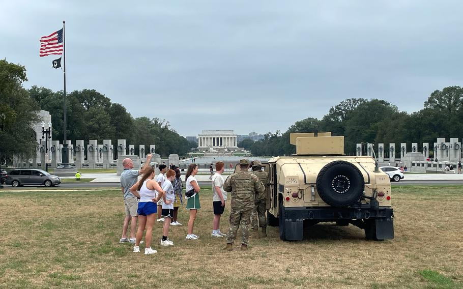 A family stands next to two soldiers by an armored vehicle, with the Lincoln Memorial and reflecting pool in the background.