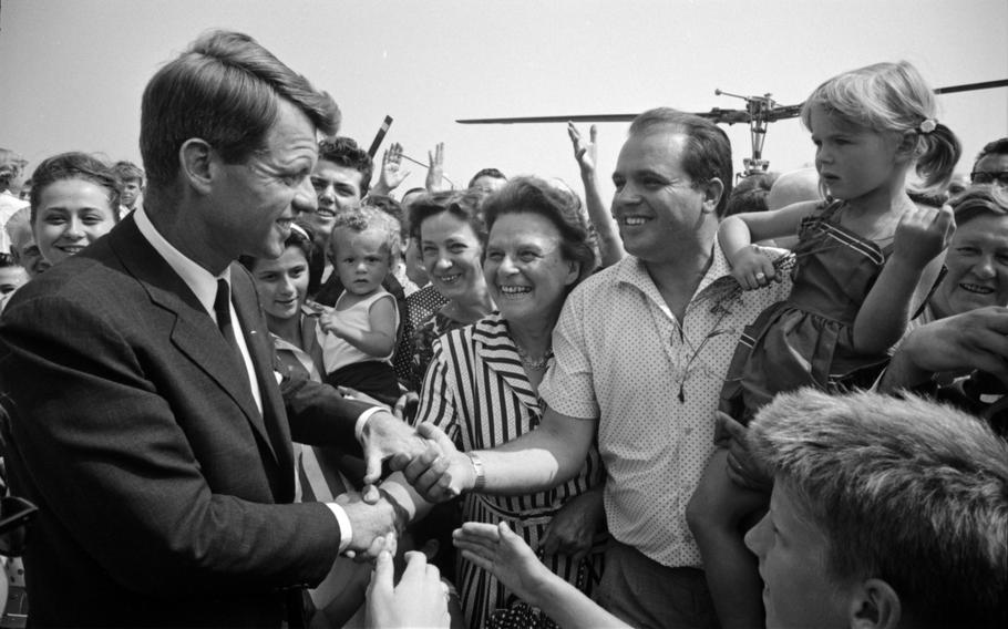 RFK shaking the hands of two people as fans surround him and a helicopter’s rotors are visible in the background.
