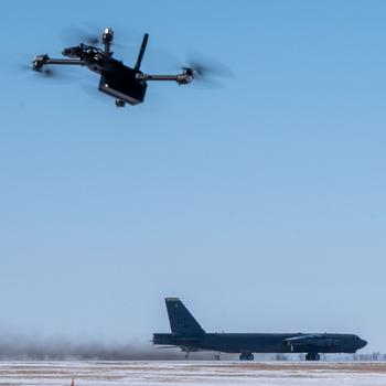 A drone flies in the foreground in the top half of the image, while a plane is on a runway in the background, in the bottom half of the image.