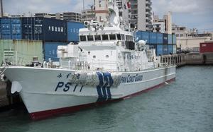 A white Japan Coast Guard patrol vessel with hull number PS11 docked at a busy commercial port. The ship displays both Japanese characters and “JAPAN COAST GUARD” in English along its hull. 