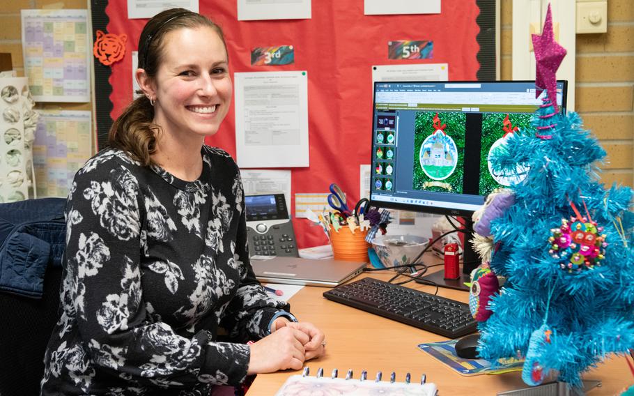 A teacher smiles while sitting at her desk, which has a computer and a blue Christmas tree.