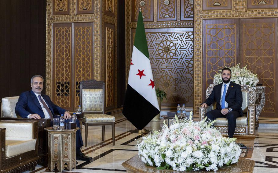 Two men in blue and black suits sit in an ornate hallway.