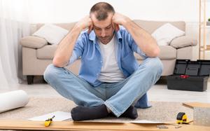 Confused Mature Man Having Hard Time Assembling Furniture Reading Self-Assembly Manual For Wooden Shelf Posing Sitting On Floor Indoor. Male Struggling Furnishing And Installing Furniture At Home