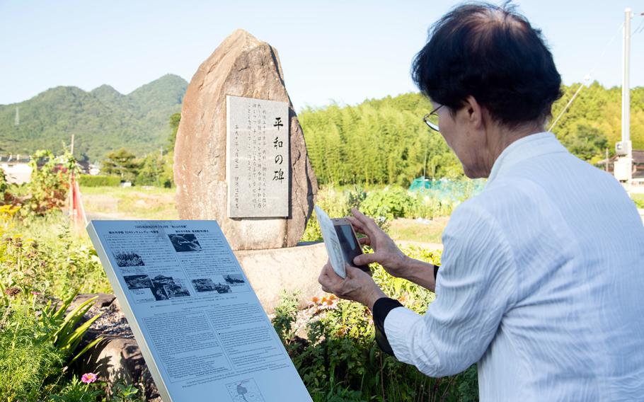 An elderly Japanese woman wearing a white shirt and glasses, seen from behind, uses a phone to take a photo of a plaque in front of a stone memorial marker, with forested hills in the background.