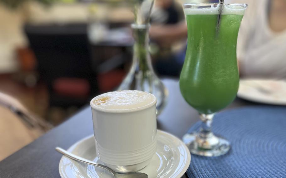 A table setting showing a white coffee cup on a saucer next to a tall glass containing a bright green beverage with a straw. 