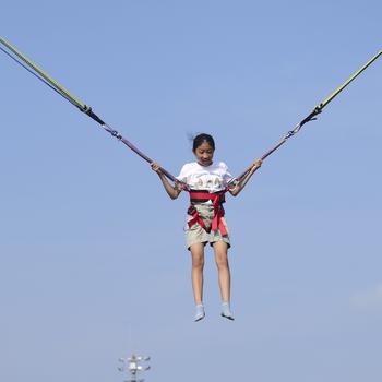 A child in shorts and T-shirt wears a harness around her waist with large bungee cords attached to either side of the harness and blue sky behind.