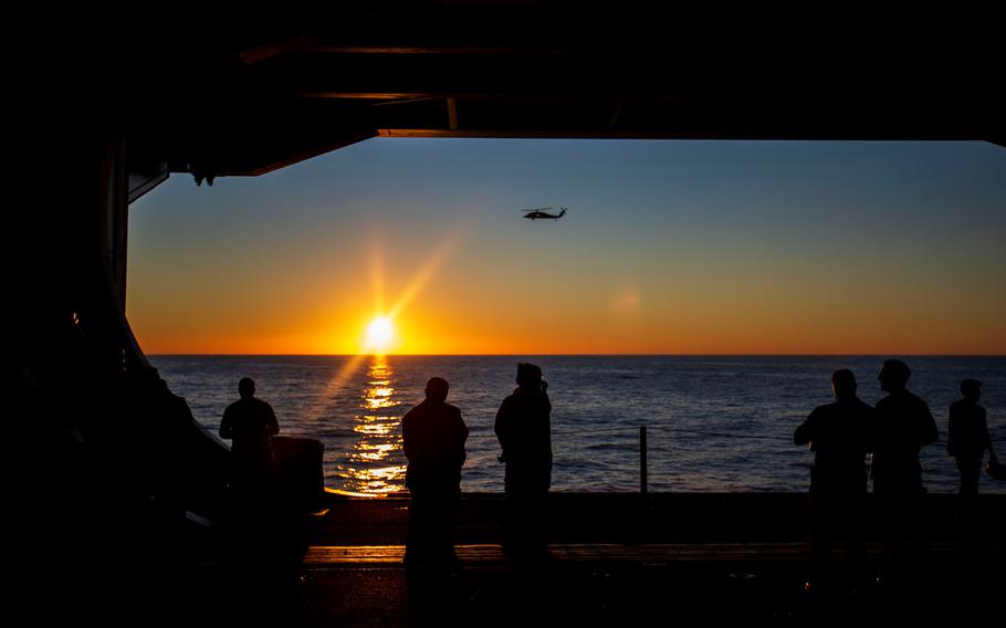 Sailors and crew members gaze out of the hangar bay at the sunset as a helicopter flies past the USS Gerald R. Ford in the Atlantic Ocean on Thursday, Oct. 6, 2022.
