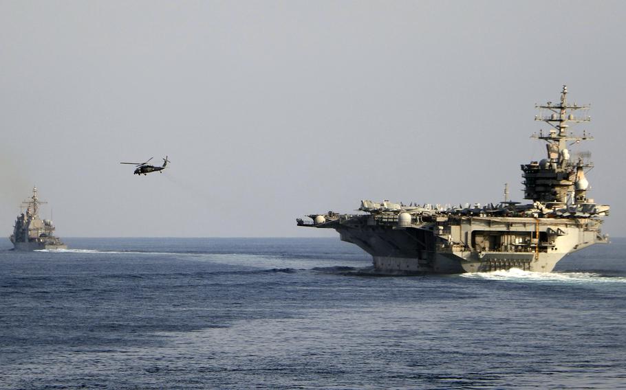 An aircraft carrier sails in open ocean, with another warship sailing on its left and a helicopter flying between them.