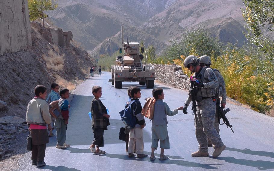 Two soldiers talk with small children on a road in a mountainous area, with an armored vehicle ahead of them on the road.
