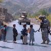 Two soldiers talk with small children on a road in a mountainous area, with an armored vehicle ahead of them on the road.
