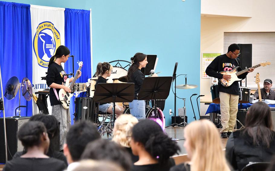 A quartet of teenagers, all wearing the same black t-shirts reading “Donuts of Infinity” perform on a stage. 