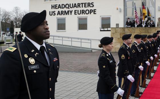 Soldiers in dress uniforms and berets stand in formation on a stone plaza outside a building with black lettering reading “Headquarters US Army Europe.”