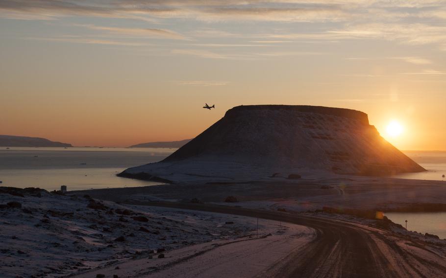 A Canadian C-130 passes Mount Dundas and approaches the runway at Thule as the sun sets over Baffin Bay. Some houses of the abandoned village are seen in the foreground.
