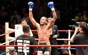 A boxer in black shorts stands in a ring and raises his arms in celebration while a trainer stands in front of him and a man with a large camera films.