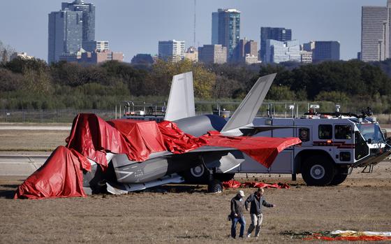 Officials look over the F-35B military aircraft wreckage, including the parachute (right) along the runway at the Naval Air Station Joint Reserve Base in Fort Worth, December 15, 2022. The pilot ejected successfully as the plane bounced off the runway after hovering. The Fort Worth skyline can be seen in the background.