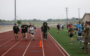 A man in a bomb suit runs on a track with several others running with him on the track and even more watching from the side.