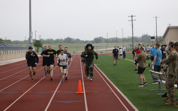 A man in a bomb suit runs on a track with several others running with him on the track and even more watching from the side.