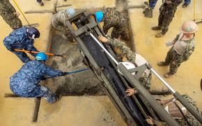 Military service members wearing camouflage uniforms and hard hats, seen from above, use construction equipment to pour cement into a large hole in asphalt.