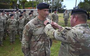 A U.S. Army ceremony where a senior officer pins onto a soldier’s uniform. The recipient, wearing multicam fatigues and a black beret, stands at attention while another soldier in similar uniform performs the pinning. 